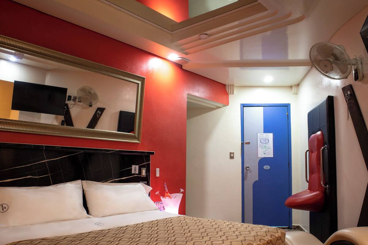 A hotel room with a bed, a mirror, a red wall, a blue door, and a fan.