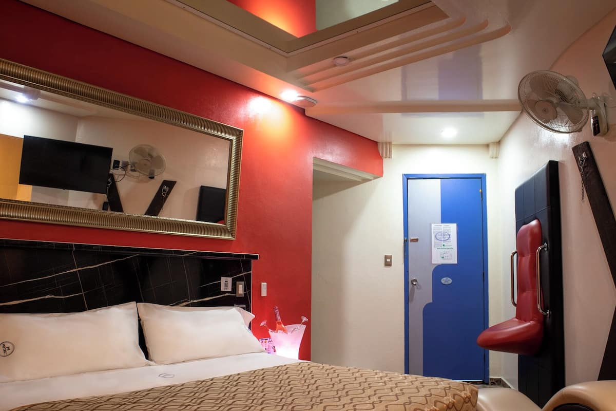 A hotel room with a bed, a mirror, a red wall, a blue door, and a fan.