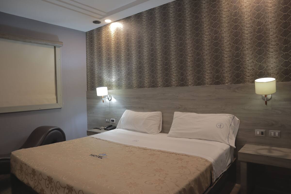 A hotel room with a bed, bedside lamps, a nightstand, a chair, and a wall-mounted TV.
