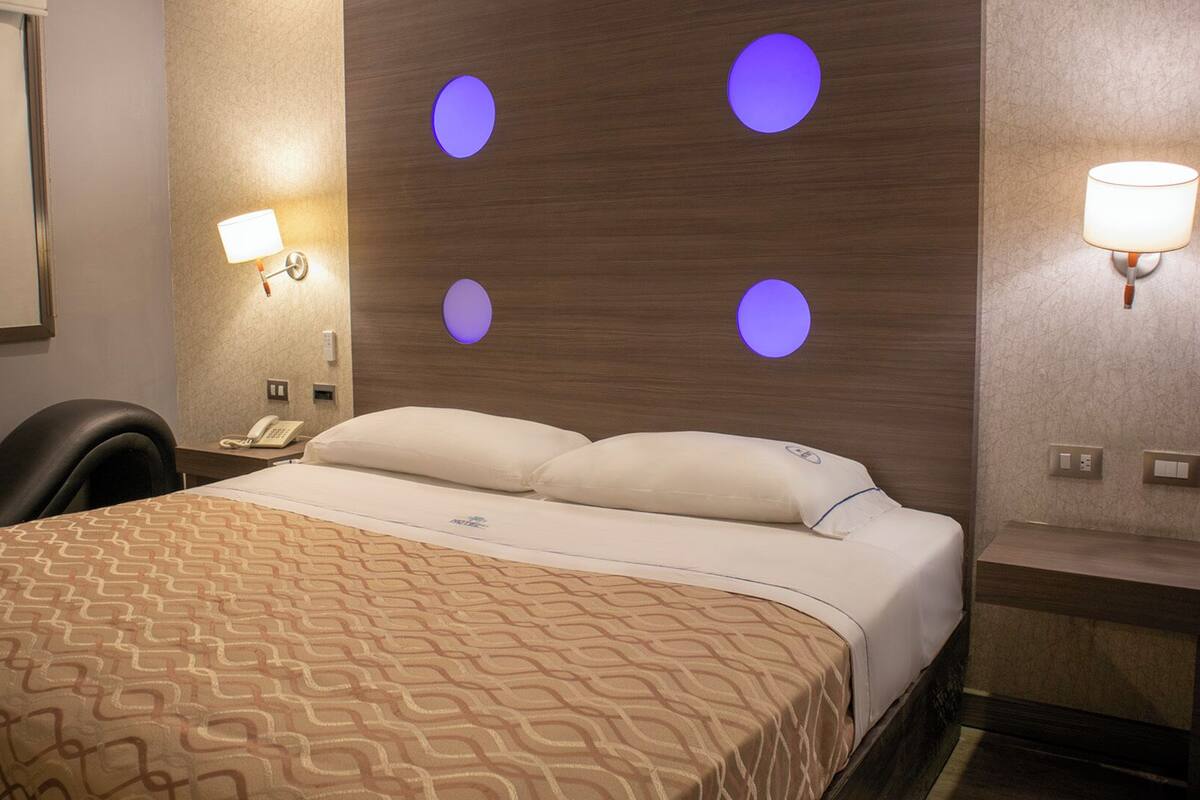 A hotel room with a bed, a desk with a lamp, and a wall-mounted phone.