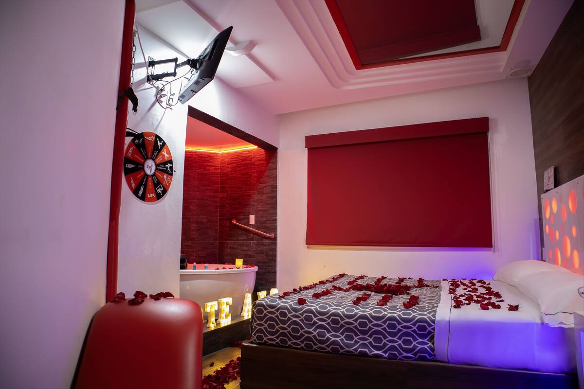 A modern bedroom with a large bed, a red accent wall, a ceiling fan, and a wall-mounted TV.