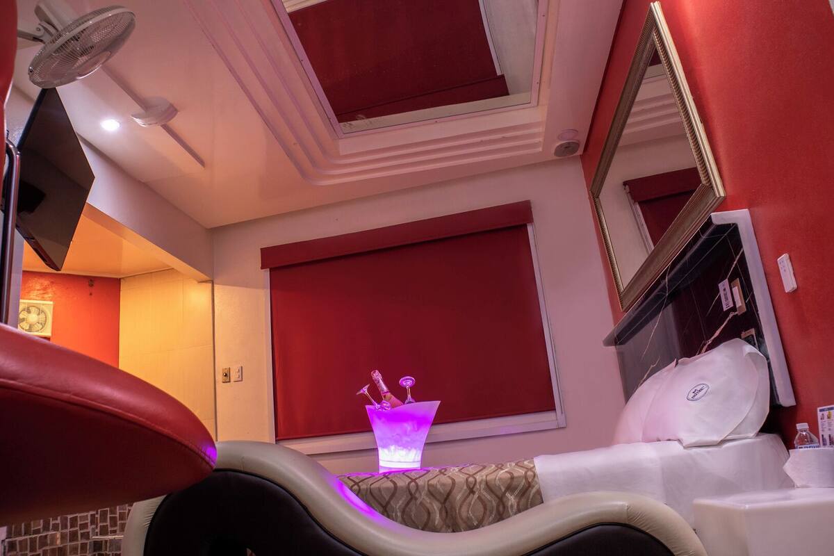 A hotel room with a red and white color scheme, a large mirror, a bed with white linens, and a small table with a purple light.