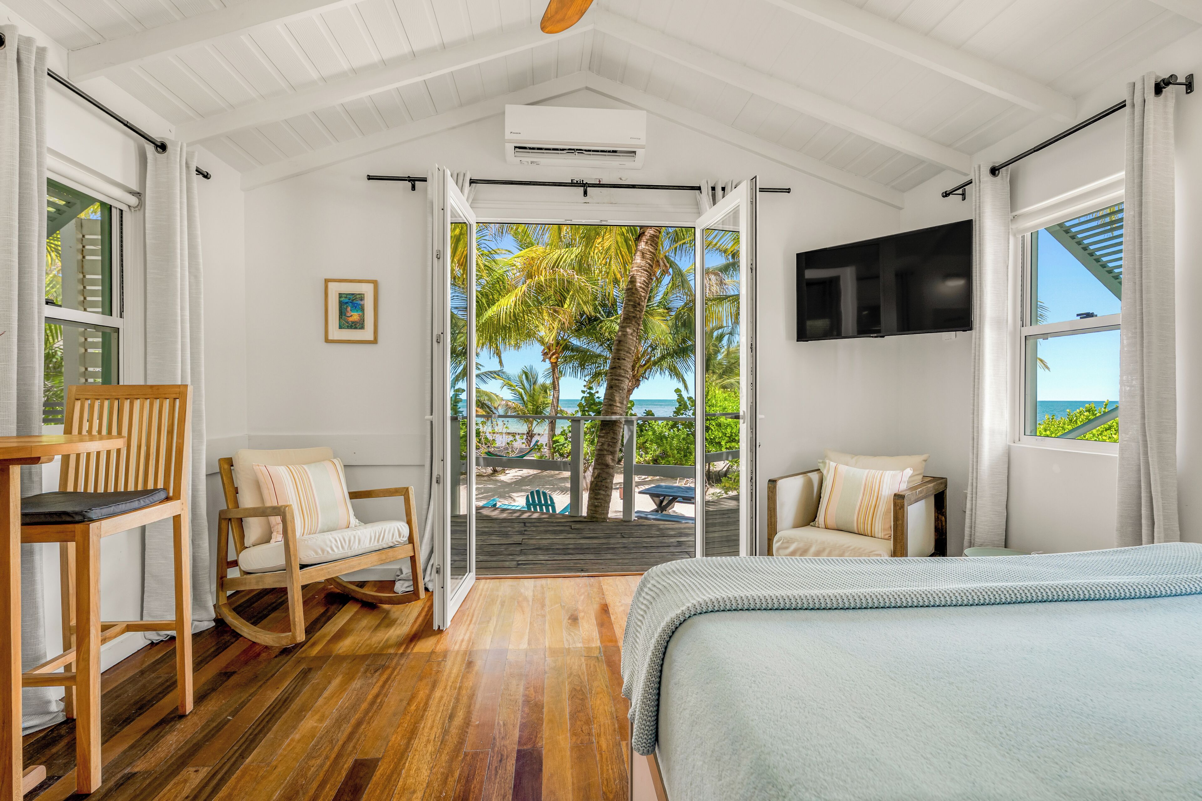 A room with a bed, a wooden chair, a rocking chair, a TV, and a view of the outdoors.