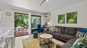 A modern living room with a grey sofa, a round coffee table, and a blue armchair. There is a dining area with white chairs and a table, and a large window with a view of greenery.