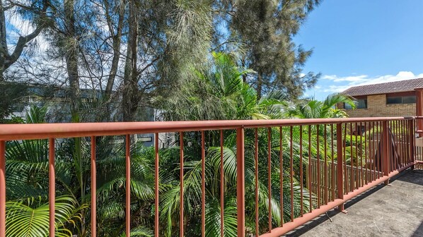 A balcony with a red metal railing, surrounded by lush green tropical plants and trees.
