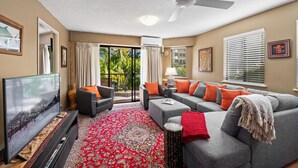 A living room with a flat-screen TV, grey sofas, red and orange cushions, a red patterned rug, and a ceiling fan. - Asiatic Suite at Nautilus Mooloolaba (Mooloolaba)