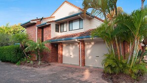 A two-story house with a garage, surrounded by tropical plants and a clear sky.