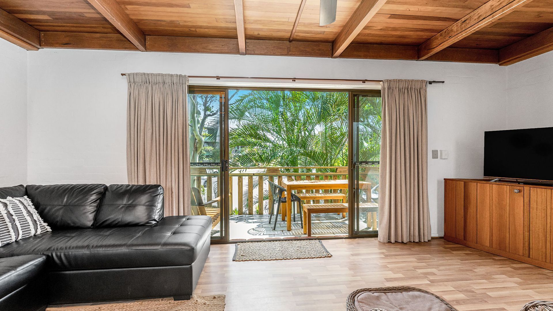 A modern living room with a black leather sofa, wooden flooring, a TV on a wooden cabinet, and a sliding glass door leading to a balcony with a wooden table and chairs.