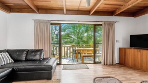 A modern living room with a black leather sofa, wooden flooring, a TV on a wooden cabinet, and a sliding glass door leading to a balcony with a wooden table and chairs.