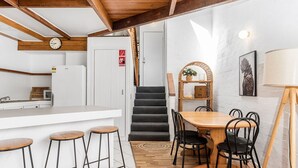 A modern kitchen with a white fridge, wooden stools, a dining table with chairs, and a staircase leading to another room.