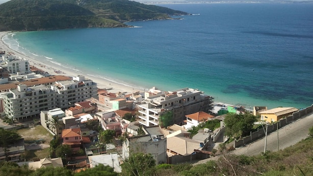 A coastal town with a beach, buildings, and a mountain in the background. - Located 150 meters from the most charming beach in Arraial do Cabo, this 2-bedroom apartment features air conditioning! (Arraial do Cabo)