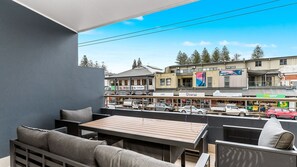 Un balcon avec des sièges pour l’extérieur, une vue sur une rue bordée de divers commerces et un ciel dégagé.