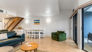 Un salon moderne comprenant un canapé, une table basse, un coin repas avec une table et des chaises, un fauteuil vert et un escalier.