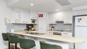Une cuisine moderne avec des meubles blancs, un îlot central doté de tabourets de bar, un four à micro-ondes, une machine à café et un panneau indiquant l’emplacement de l’extincteur.