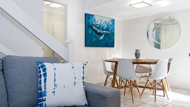 Ett modernt vardagsrum med en grå soffa, en blå och vit kudde, ett matbord med vita stolar, en blå målning av en val och en rund spegel på väggen.