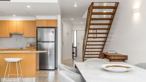 A modern kitchen with wooden cabinets, a stainless steel refrigerator, and a dining area with a marble table and white chairs. - Byron Bay Accom - Solace 1 - 8 Lawson Street (Byron Shire)
