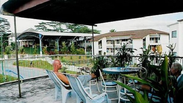 A covered outdoor seating area with chairs and a table, surrounded by plants and a view of a building with a sloped roof.