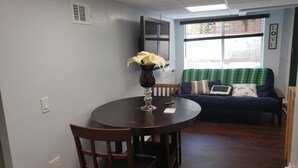 A dining area with a round table, chairs, a vase with flowers, a window with blinds, and a sofa with cushions.