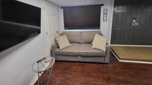 A living room with a grey sofa, two patterned cushions, a small glass table with a remote control, and a dark door with a 'LOVE' sign.