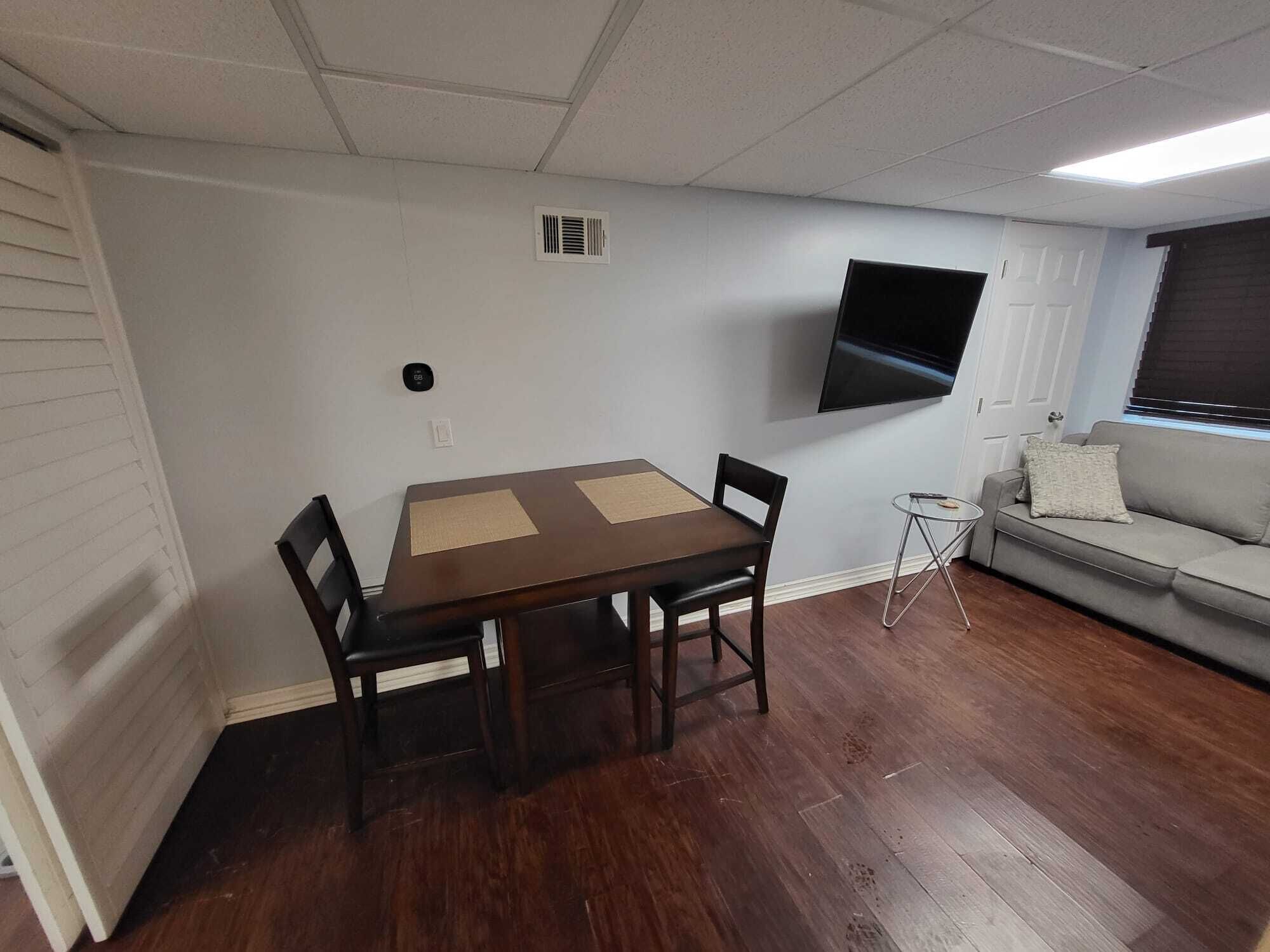 A room with a wooden dining table, two chairs, a sofa, a TV mounted on the wall, and a small side table.