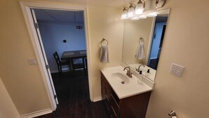 A bathroom with a white sink, a mirror, and a towel rack. There is a dark wooden floor and a door leading to a dining area.