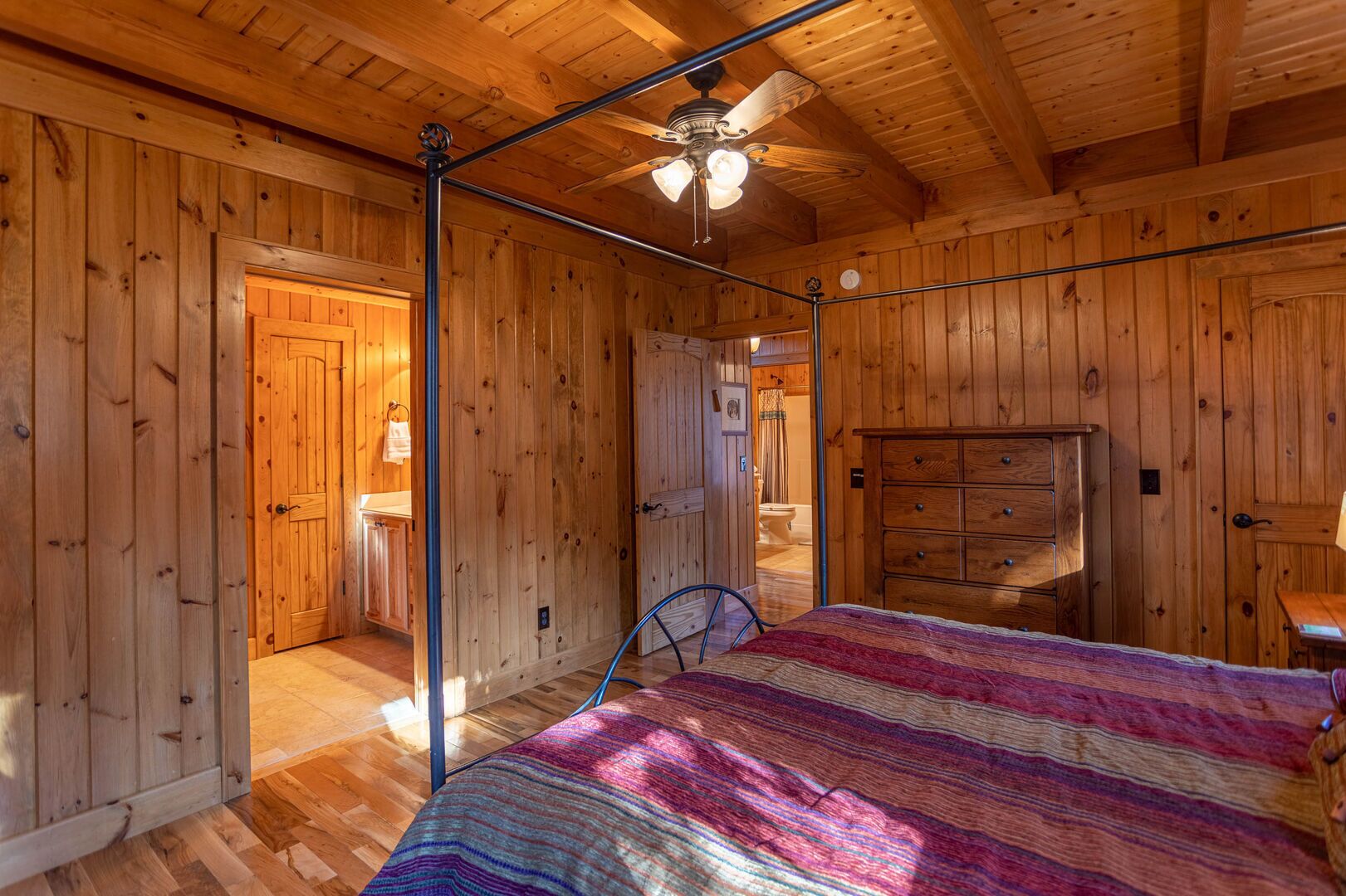 A wooden cabin bedroom with a bed, a dresser, a ceiling fan, and a bathroom visible through an open door.