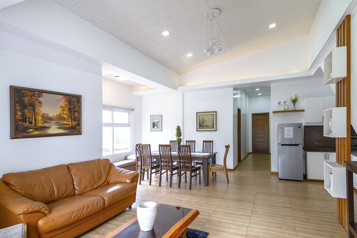 A spacious living area with a leather sofa, dining table, and chairs, a kitchen with a refrigerator, and a painting on the wall.