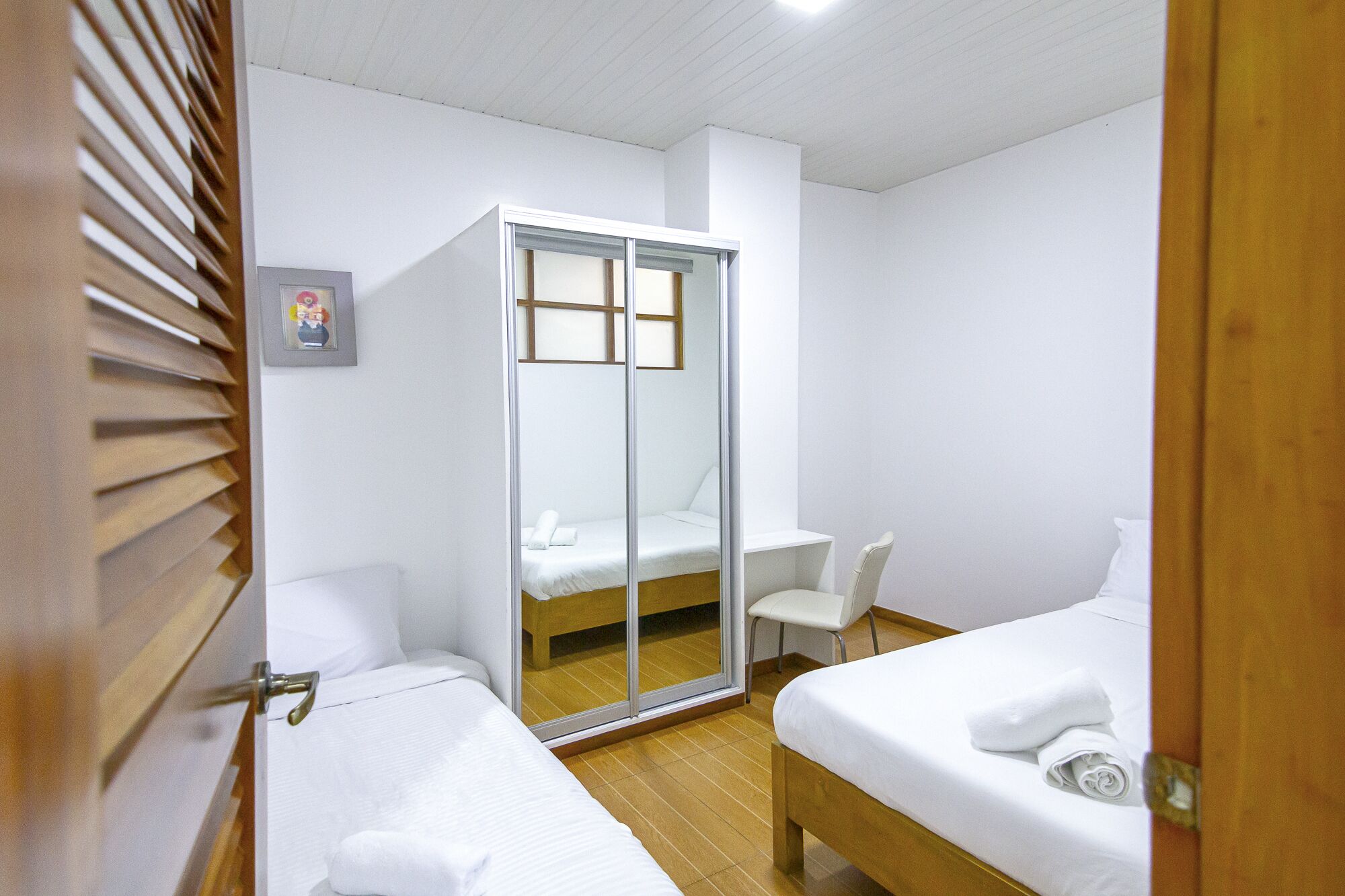 Quarto de hotel com duas camas, piso de madeira, parede branca, porta de madeira e uma cadeira branca pequena.