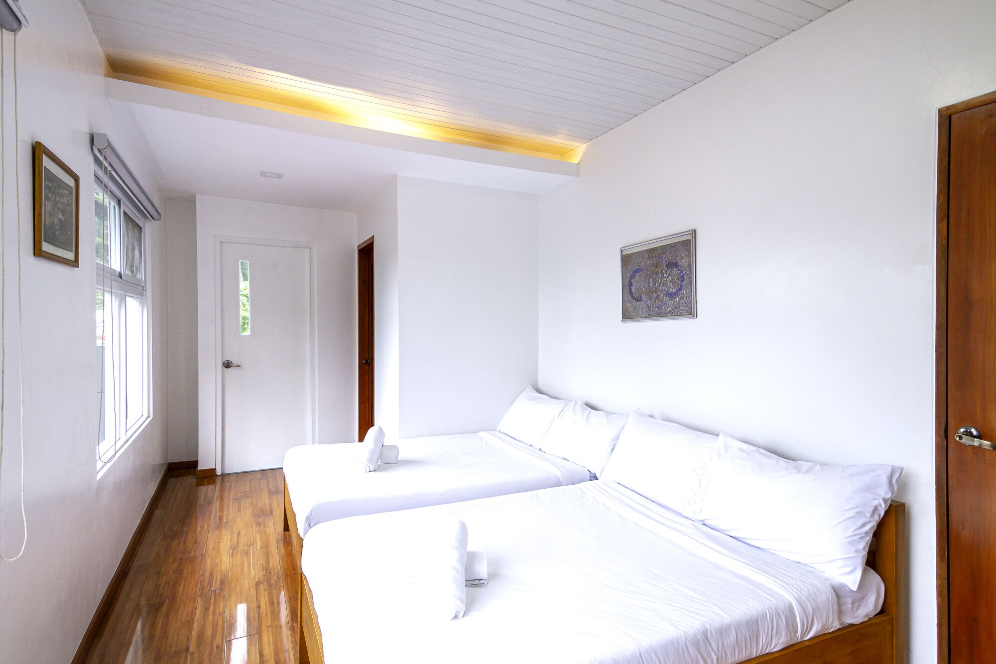 A room with two beds, wooden flooring, a white door, and a framed picture on the wall.