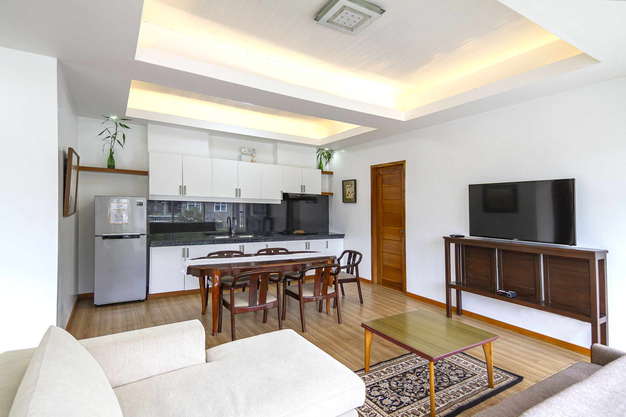 Una sala de estar moderna con un sofá, una mesa de comedor con sillas, una zona de cocina con armarios y electrodomésticos, y un televisor sobre un mueble de madera.