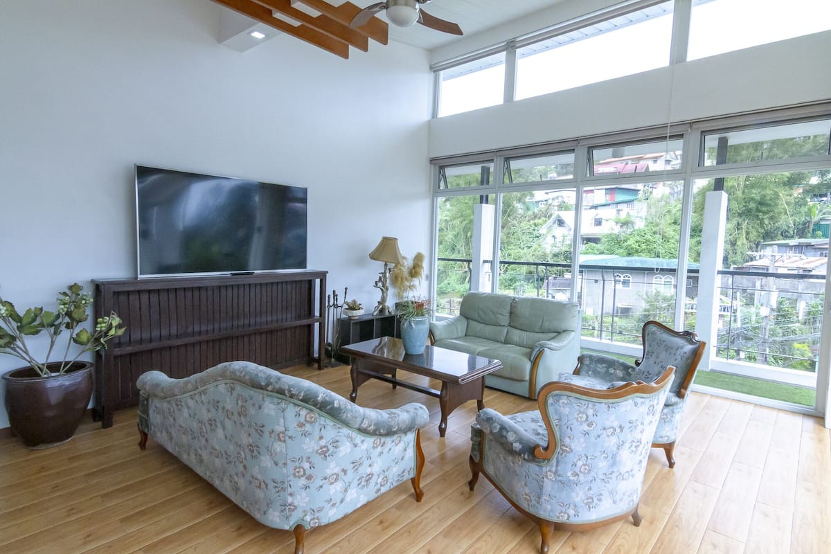 A spacious living room with a large TV, floral-patterned furniture, a wooden coffee table, and a view of a cityscape through large windows.