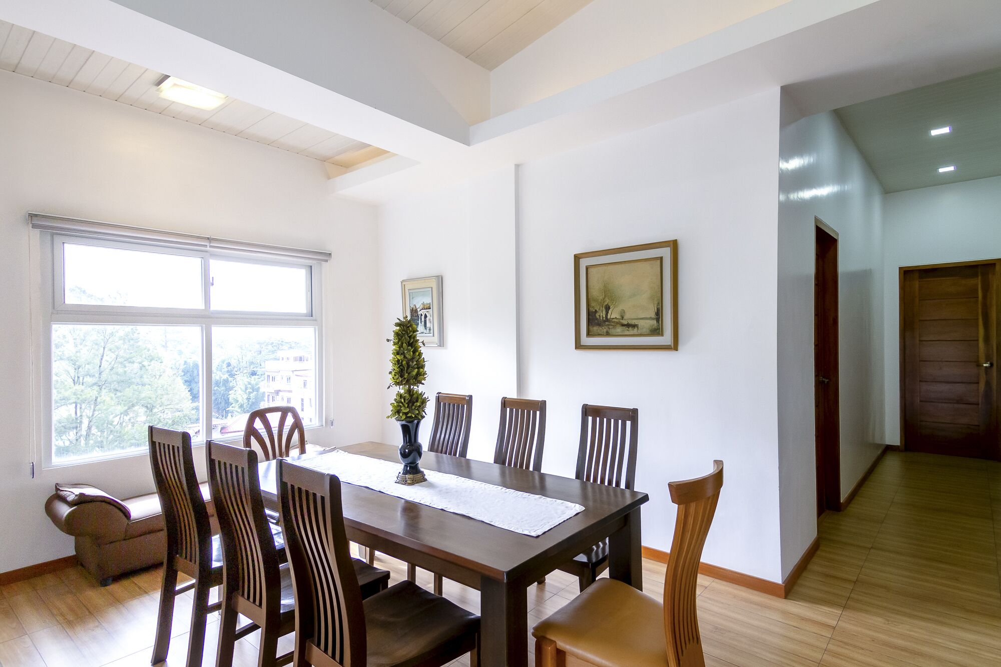 Uma sala de jantar com uma mesa e cadeiras de madeira, um vaso com uma planta e um quadro na parede.