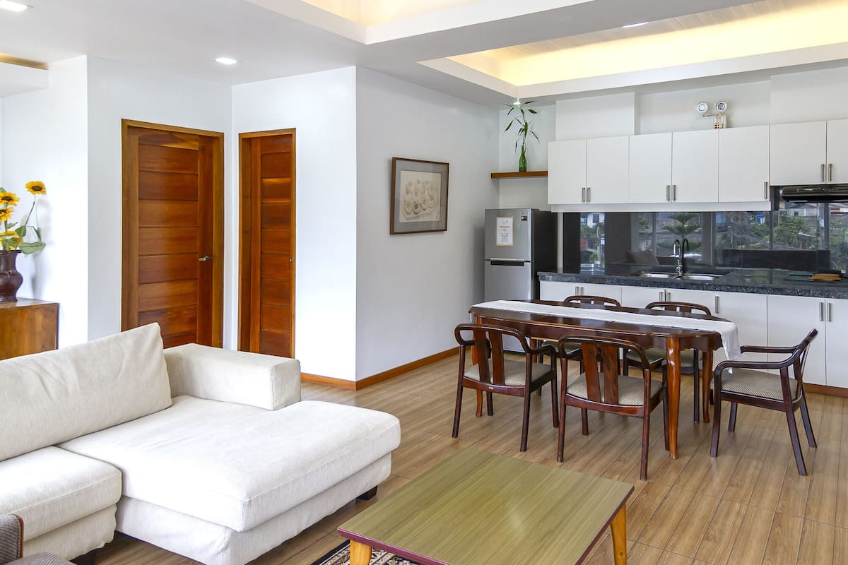 A modern living room with a sofa, dining table, and chairs. There is a kitchen area with cabinets and a refrigerator.
