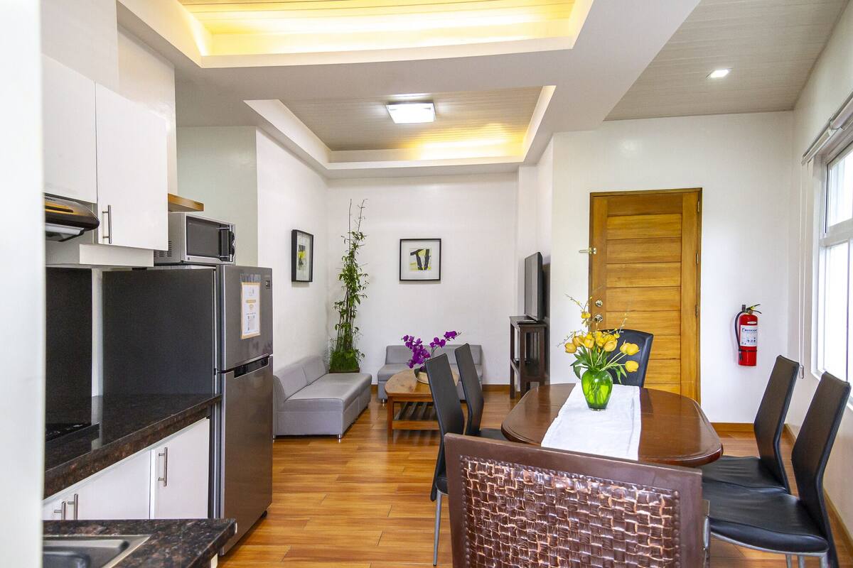 A modern kitchen with a refrigerator, microwave, and stove. There is a dining area with a table, chairs, and a vase with flowers. A television is mounted on the wall, and a wooden door is visible in the background.