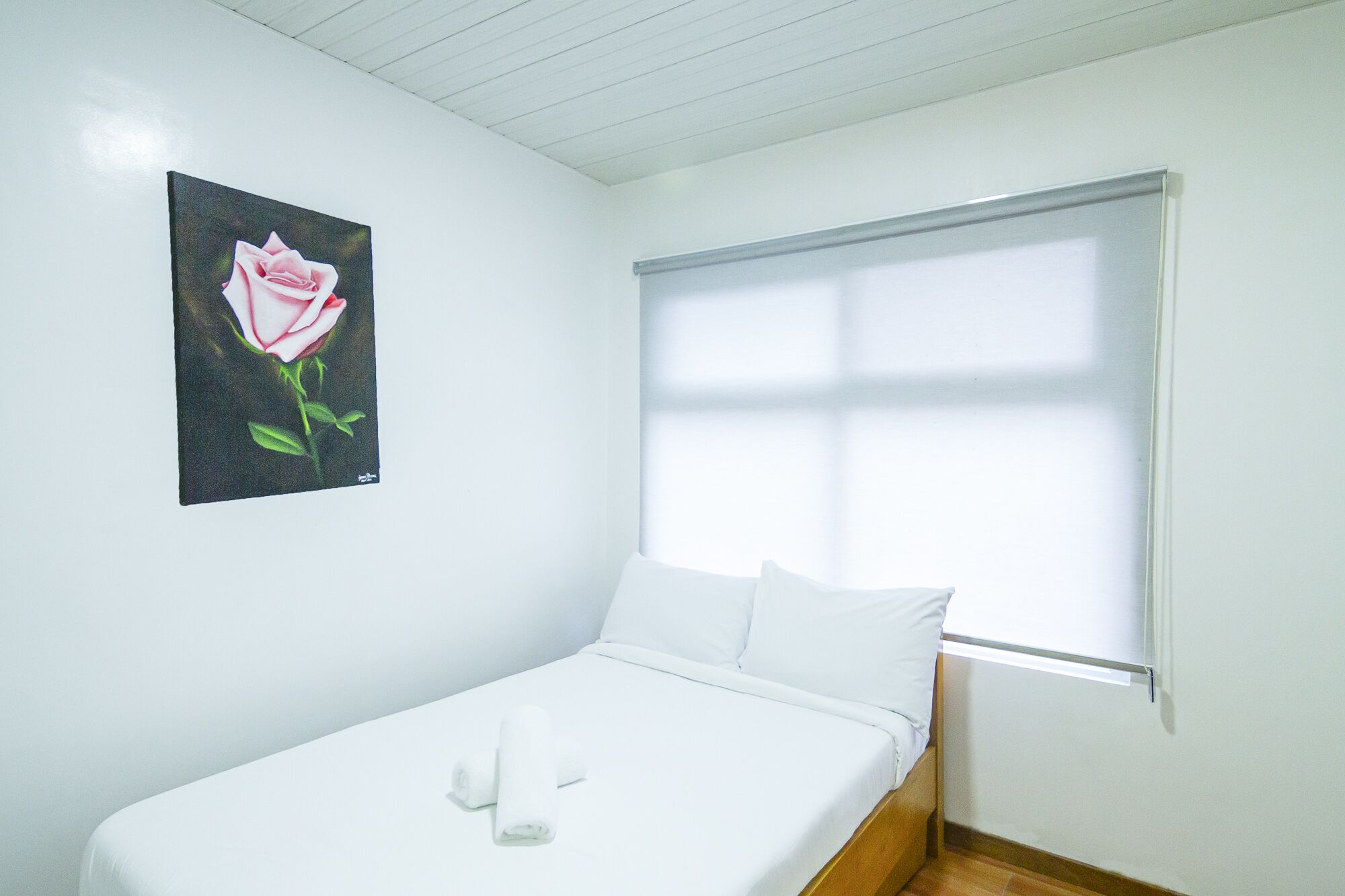 Een slaapkamer met een enkel bed, wit beddengoed, een raam met zonwering en een schilderij van een roos aan de muur.