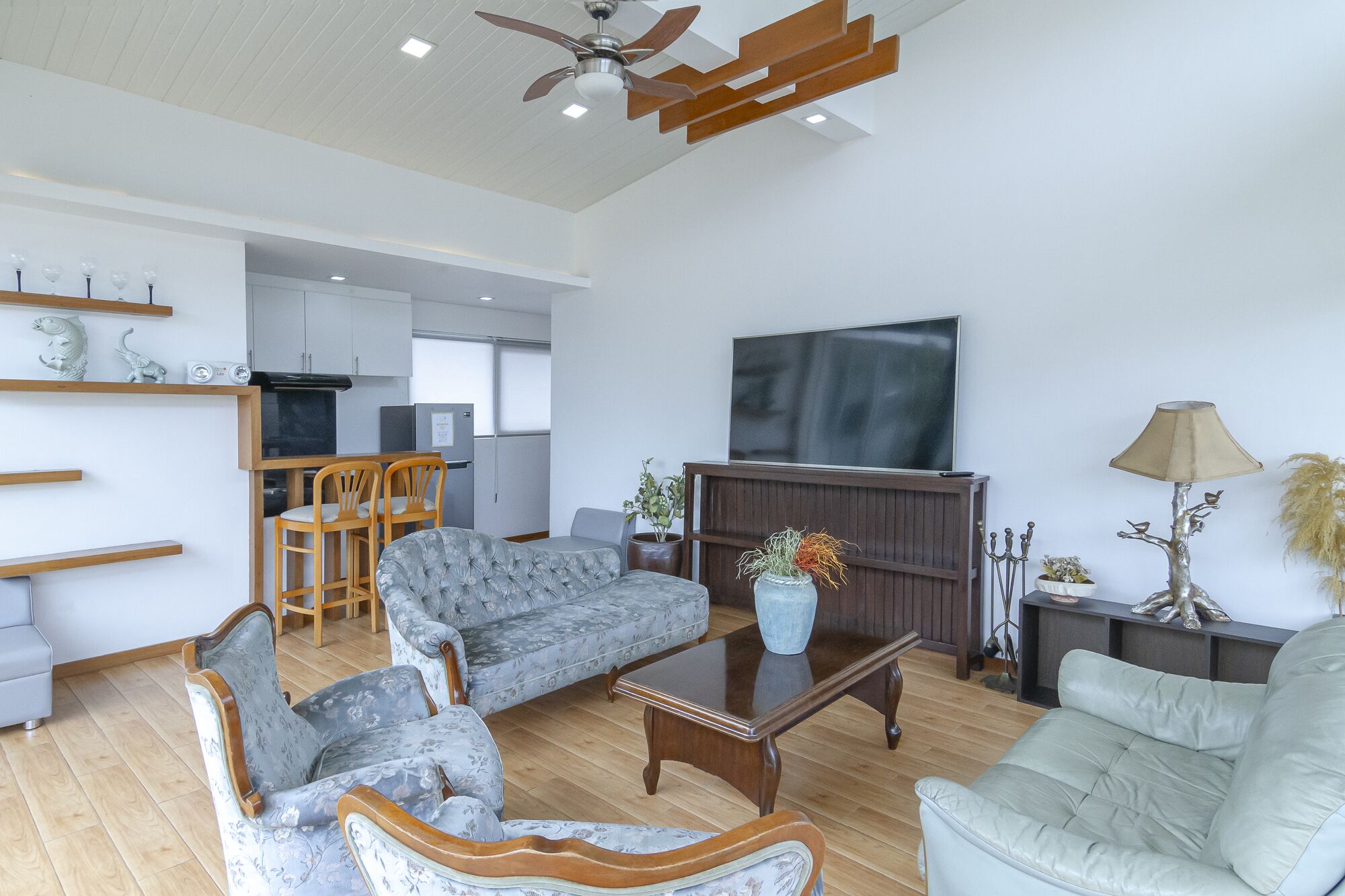 A spacious living room with a sofa, armchairs, a coffee table, and a television. There is a kitchen area in the background with a dining table and chairs.