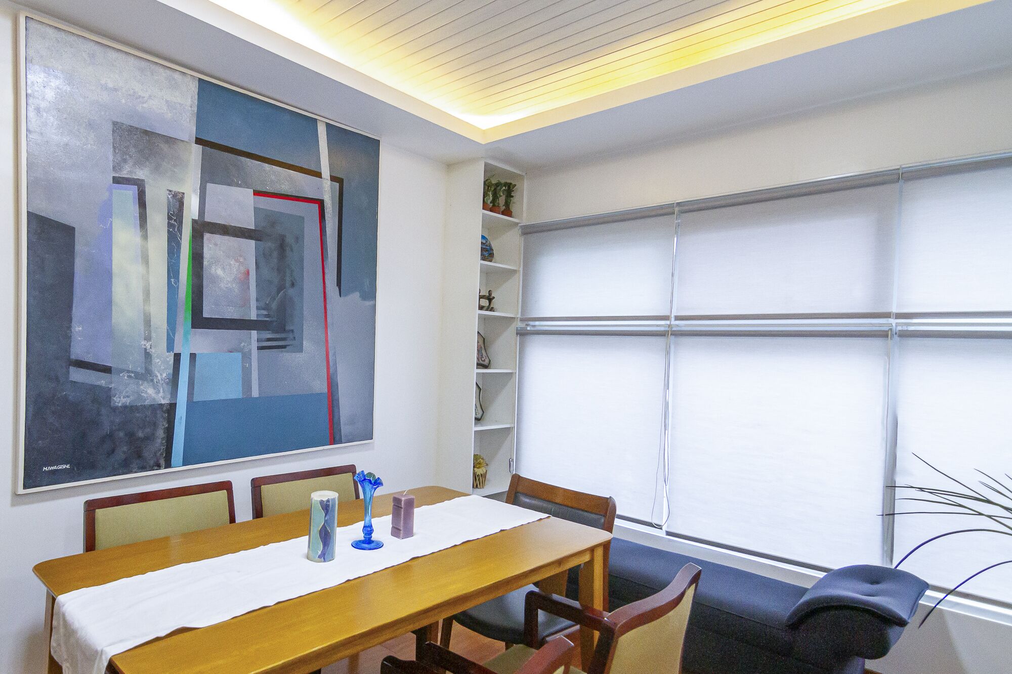 Una habitación con un cuadro abstracto grande, una mesa de madera con sillas, una ventana con persianas y una estantería con objetos decorativos.