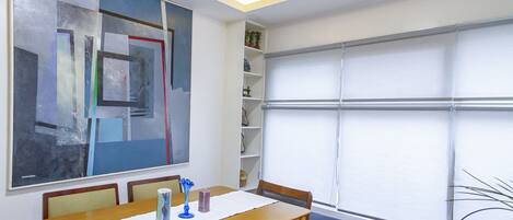 Una habitación con un cuadro abstracto grande, una mesa de madera con sillas, una ventana con persianas y una estantería con objetos decorativos.