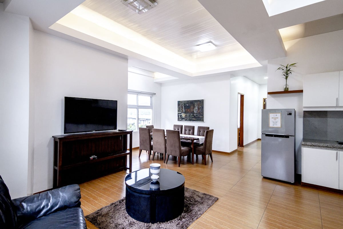 A modern living room with a flat-screen TV, a dining area with a table and chairs, and a kitchen with a refrigerator and a plant.