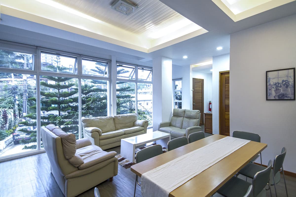 A modern living room with a sofa, armchairs, and a dining table. Large windows offer a view of a garden and trees.