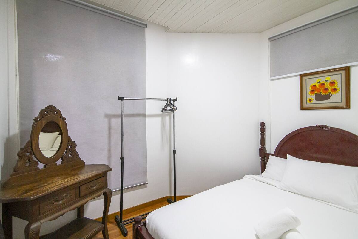 A bedroom with a wooden dresser, a mirror, a bed with white bedding, a wooden headboard, and a framed picture on the wall.