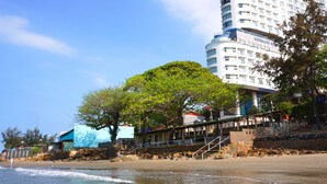 Un hotel frente a la playa con un gran árbol en primer plano y una escalera que lleva a la entrada, con vista al océano.