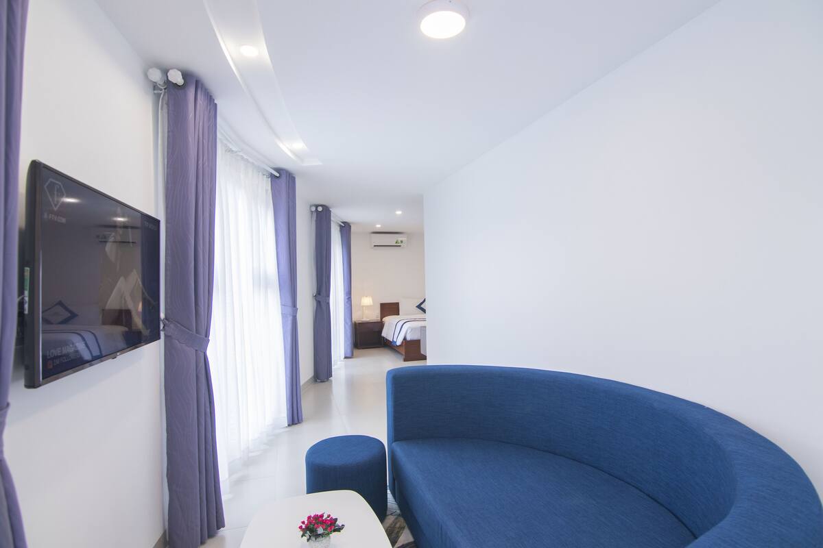 A modern hotel room with a curved blue sofa, a flat-screen TV mounted on the wall, and a small table with a vase of flowers.