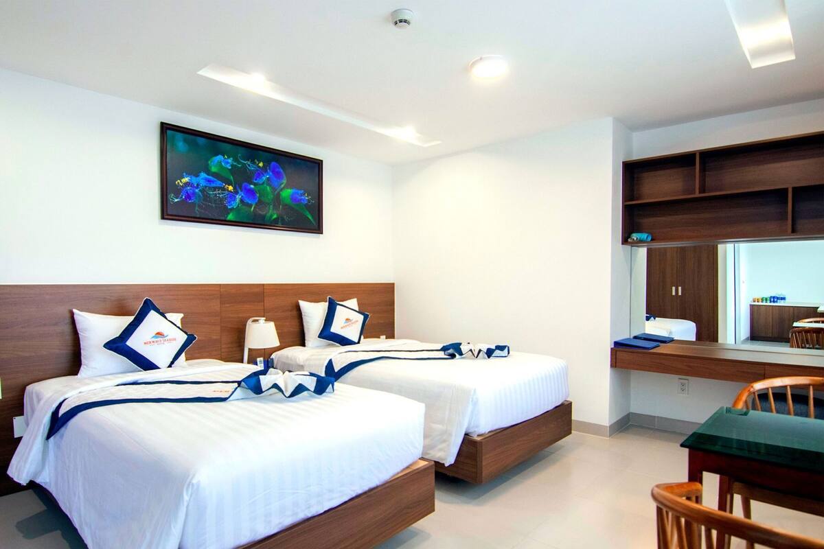 A hotel room with two beds, a desk, a chair, a painting on the wall, and built-in shelves.