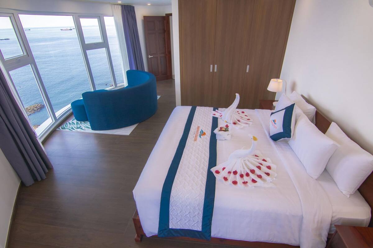 A hotel room with a bed, a blue chair, a view of the sea, and a wooden wardrobe.