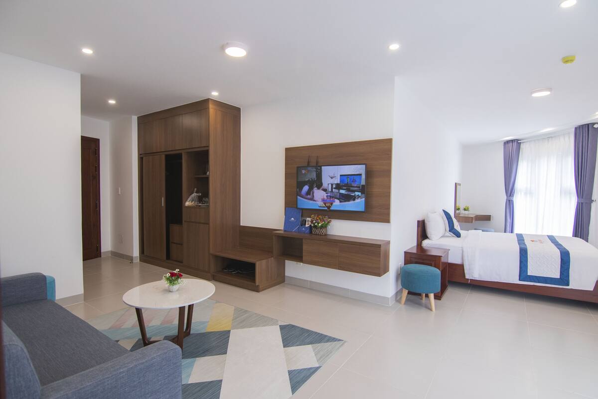 A modern hotel room with a sofa, a small round table with flowers, a TV mounted on the wall, a bed with a blue and white bedspread, and a wardrobe.