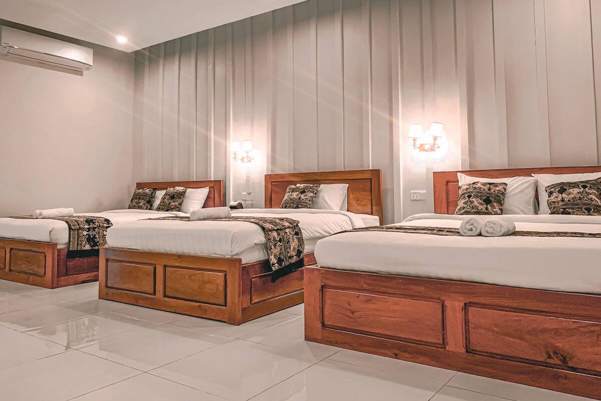 A hotel room with three wooden beds, each with white linens and patterned pillows. There are wall-mounted lamps on either side of the beds.