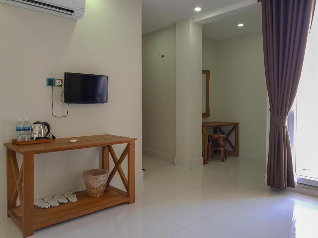 A hotel room with a wooden console table, a television mounted on the wall, a small table with a chair, and a window with curtains.
