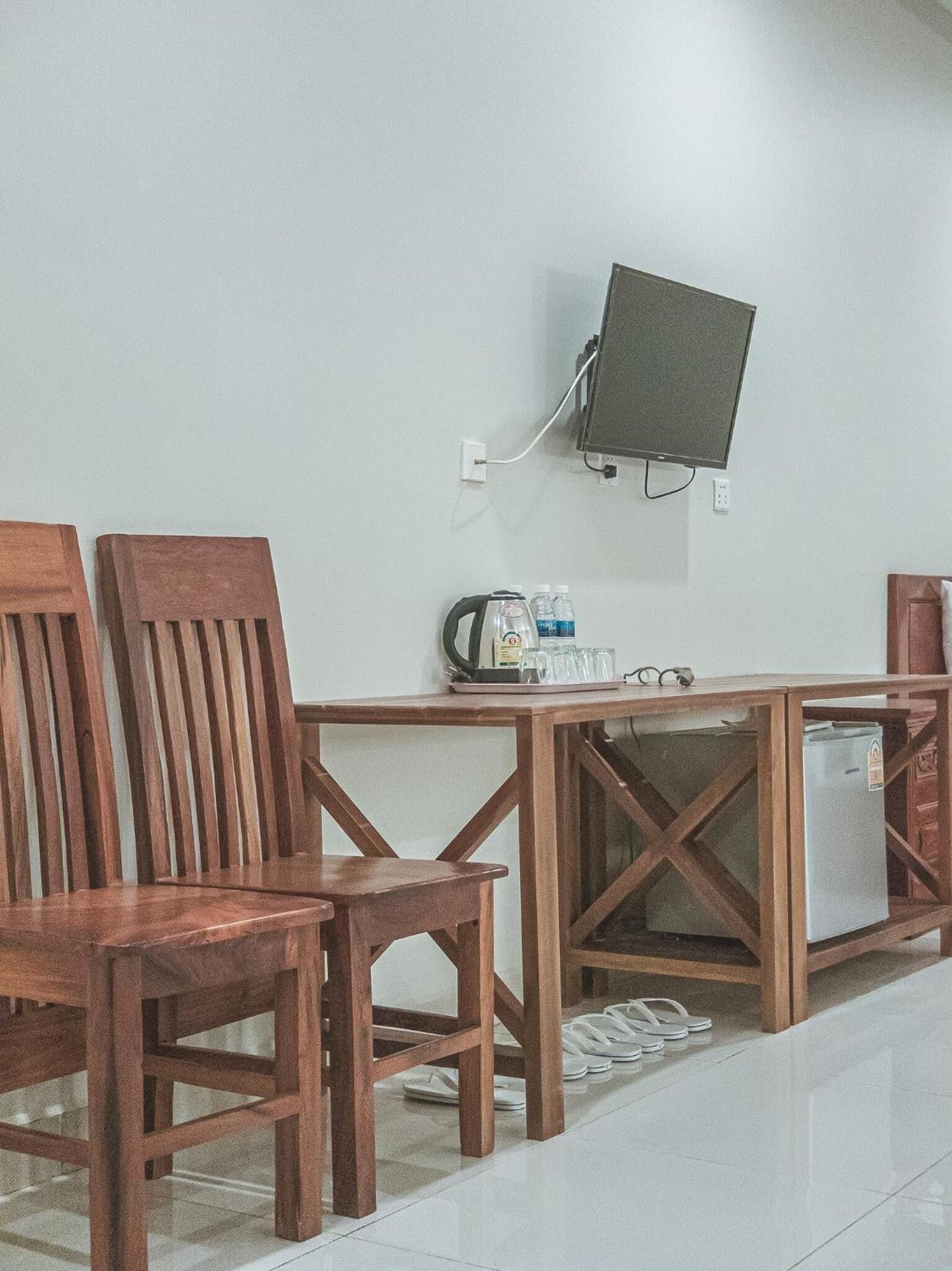 Ein Zimmer mit zwei Holzsesseln, einem kleinen Tisch mit Wasserkocher, Gläsern und Flaschen, einem an der Wand montierten Fernseher und einem Kühlschrank.