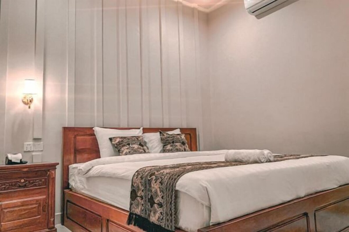 A neatly made bed with a wooden headboard, a bedside table with a lamp, and a wall-mounted air conditioner.